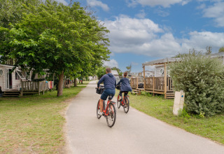 Two people ride bicycles at Flower Camping Les Paludiers, passing mobile homes and lush green trees.