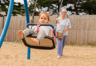 A child swings at the playground of Flower Camping Les Paludiers, watched by an elderly woman in the background.