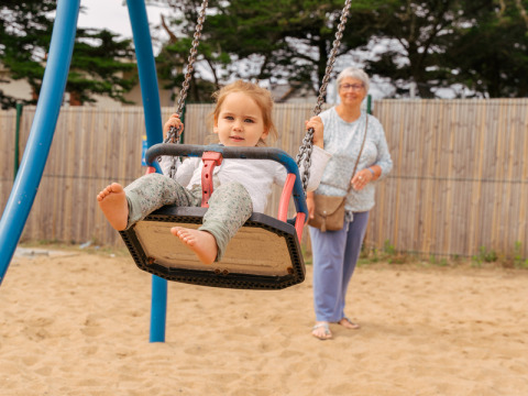 A child swings at the playground of Flower Camping Les Paludiers, watched by an elderly woman in the background.