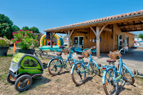Bicicletas y remolque infantil junto a la recepción y zona de juegos en Flower Camping Le Bel Air, Francia.