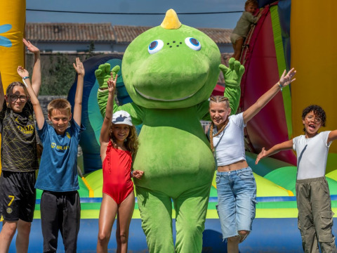 Niños sonríen junto a una mascota dinosaurio verde en un inflable en Flower Camping Le Bel Air, Francia.
