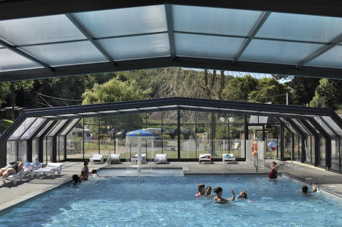 Indoor pool with glass roof and walls at Flower Camping La Chênaie in Normandy, France, with people swimming.