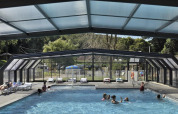 Indendørs pool med glasloft og glasvægge på Flower Camping La Chênaie i Normandiet, Frankrig, med folk der svømmer.