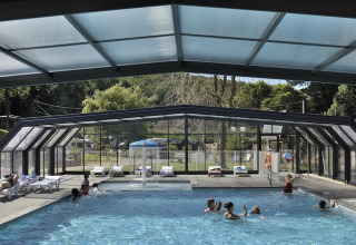Indendørs pool med glasloft og glasvægge på Flower Camping La Chênaie i Normandiet, Frankrig, med folk der svømmer.