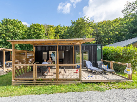Visitors relaxing on a wooden terrace outside a cabin at Flower Camping La Chênaie in Normandy, France.