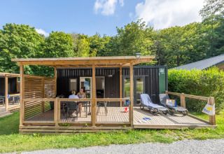 Gäste entspannen auf einer Holzterrasse vor einer Hütte im Flower Camping La Chênaie in der Normandie, Frankreich.