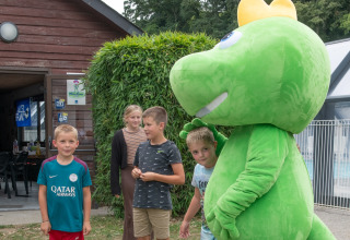 Des enfants posent avec une grande mascotte verte à Flower Camping La Chênaie, un parc de vacances en Normandie.