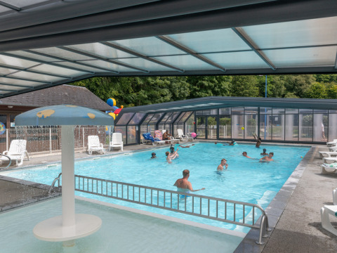 Überdachtes Schwimmbad mit badenden Gästen und Liegestühlen im Flower Camping La Chênaie, Normandie, Frankreich.