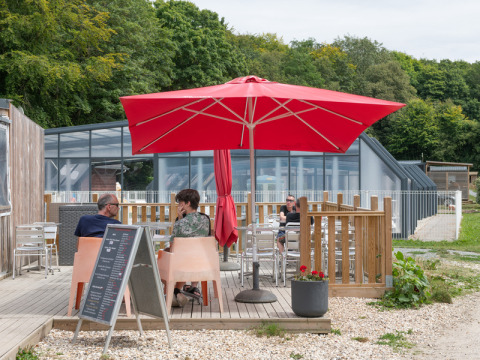 Buitenterras Flower Camping La Chênaie, Normandië, met rood parasol en gasten rond tafels.