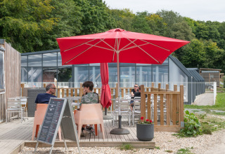 Terraza exterior en Flower Camping La Chênaie en Normandía, con sombrilla roja y varias personas sentadas.