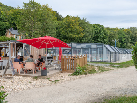 Outdoor terrace with red umbrella and people beside an indoor pool at Flower Camping La Chênaie, Normandy, France.