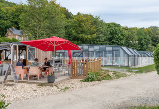Udendørs terasse med folk, rød parasol og indendørs swimmingpool ved Flower Camping La Chênaie i Normandiet.