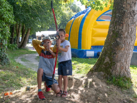 Two children play with a bow and arrow in a green park next to a large yellow and blue inflatable structure.
