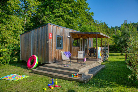 Chalet en bois avec terrasse au Flower Camping La Sagne, entouré de verdure et de jouets d’enfants sur la pelouse.