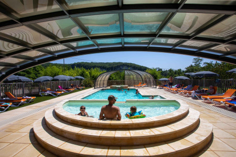 Udendørs swimmingpool med familie og solstole under overdækket område i Flower Camping La Sagne, Frankrig.