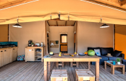 Binnenkant van een safaritent met houten meubels, keuken, zetel en eettafel op een houten vloer.