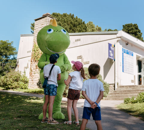 Children greeting a large green mascot outside at a holiday park in Brittany, France, with buildings behind.