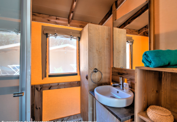 Salle de bain d’une tente safari avec lavabo, miroir, étagère, serviette roulée et lumière naturelle.