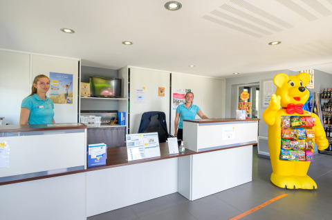 Recepción de Flower Camping Le Conleau en Bretaña, Francia, con personal y figura de oso grande.