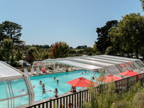 Piscina esterna coperta con telo trasparente, ombrelloni rossi e persone in un campeggio in Bretagna, Francia.