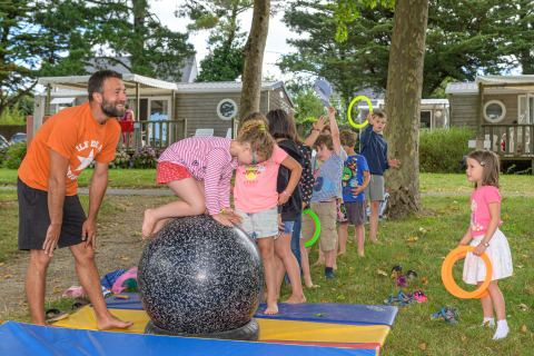 Niños juegan y se equilibran sobre una bola grande en el parque vacacional Flower Camping Le Conleau, supervisados por un adulto.