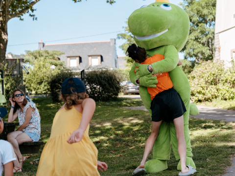 Kinderen spelen buiten, één knuffelt een grote groene mascotte op Flower Camping Le Conleau in Bretagne.