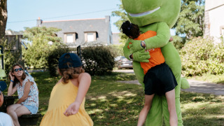 Børn leger udendørs, én krammer en stor, grøn maskot på Flower Camping Le Conleau i Bretagne, Frankrig.