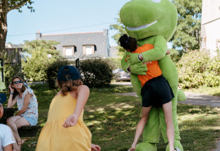 Los niños juegan al aire libre, uno abraza a una gran mascota verde en Flower Camping Le Conleau, Bretaña.