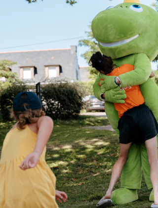 Børn leger udendørs, én krammer en stor, grøn maskot på Flower Camping Le Conleau i Bretagne, Frankrig.