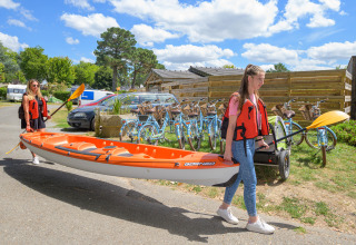Dos mujeres llevan un kayak naranja junto a bicicletas en Flower Camping Le Conleau, Bretaña, Francia.