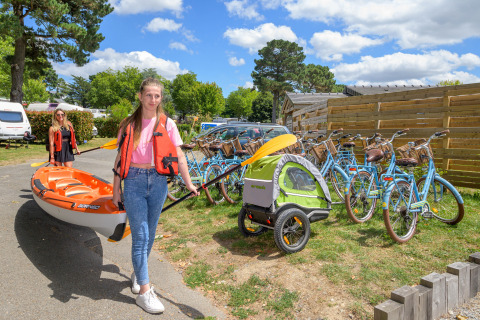 Twee vrouwen dragen een kajak naast fietsen en een aanhanger op een vakantiepark in Bretagne, Frankrijk.