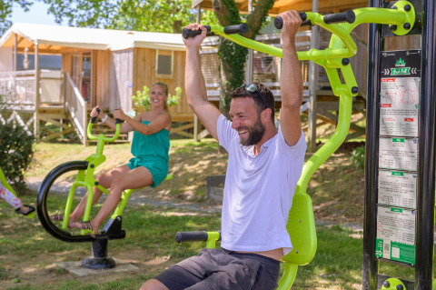 Two people exercising on outdoor fitness equipment at a holiday park in Brittany, France, on a sunny day.