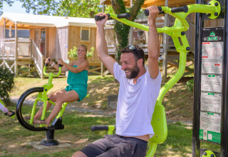 Two people exercising on outdoor fitness equipment at a holiday park in Brittany, France, on a sunny day.