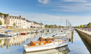 Både og sejlbåde i en idyllisk havn med smukke huse i baggrunden under en klar blå himmel.
