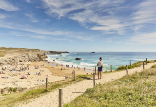 View of a sandy beach with people by the sea, seen from a pathway at a holiday park offering glamping.