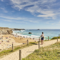 Udsigt over en sandstrand fyldt med folk ved havet, set fra en sti ved et feriepark med glamping.