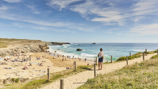 Udsigt over en sandstrand fyldt med folk ved havet, set fra en sti ved et feriepark med glamping.