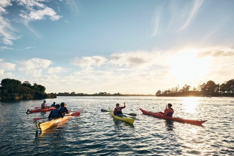 Un groupe fait du kayak au coucher du soleil sur un lac près d’un parc de glamping en pleine nature.