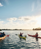 En gruppe mennesker sejler i kajak på en sø ved solnedgang nær et glampingferiecenter i naturen.