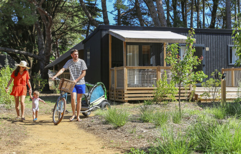 Gezin op glampingvakantie in het bos, met vader die een fiets bij een moderne houten hut voortduwt.