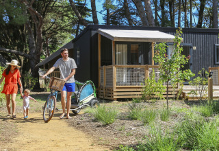 Gezin op glampingvakantie in het bos, met vader die een fiets bij een moderne houten hut voortduwt.