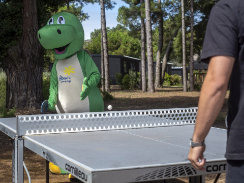 Persoon in dinosauruspak van Flower Camping Tamarins Plage speelt buiten tafeltennis in een bosrijke omgeving.