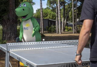 Persoon in dinosauruspak van Flower Camping Tamarins Plage speelt buiten tafeltennis in een bosrijke omgeving.