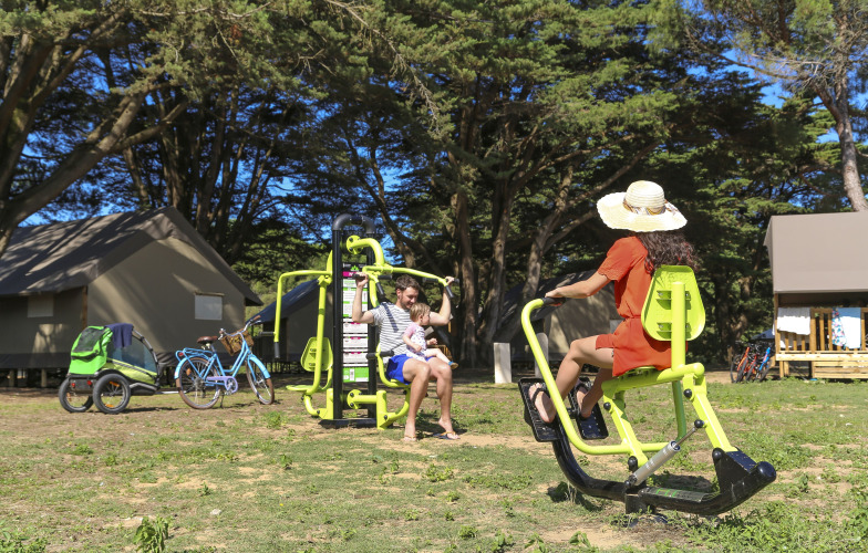 Familie nyder udendørs fitnessudstyr i solen ved telte og cykler på Flower Camping Tamarins Plage.