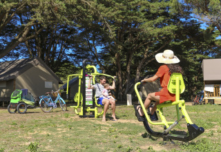 Familie beim Training an Outdoor-Fitnessgeräten auf dem Campingplatz mit Zelten und Fahrrädern in Frankreich.
