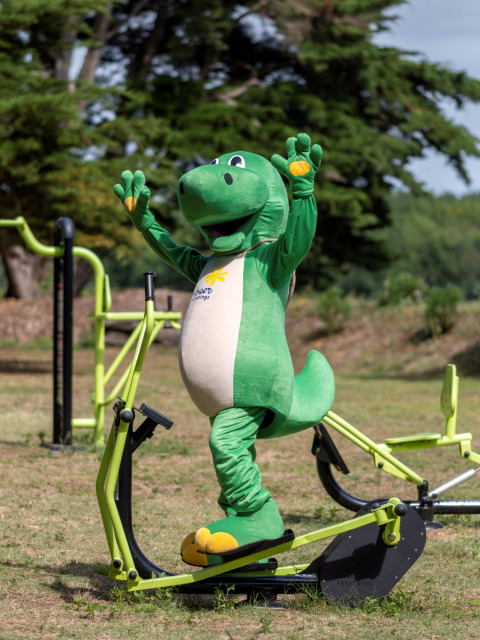Eine Person im grünen Dinosaurierkostüm benutzt ein Outdoor-Fitnessgerät im Flower Camping Tamarins Plage.