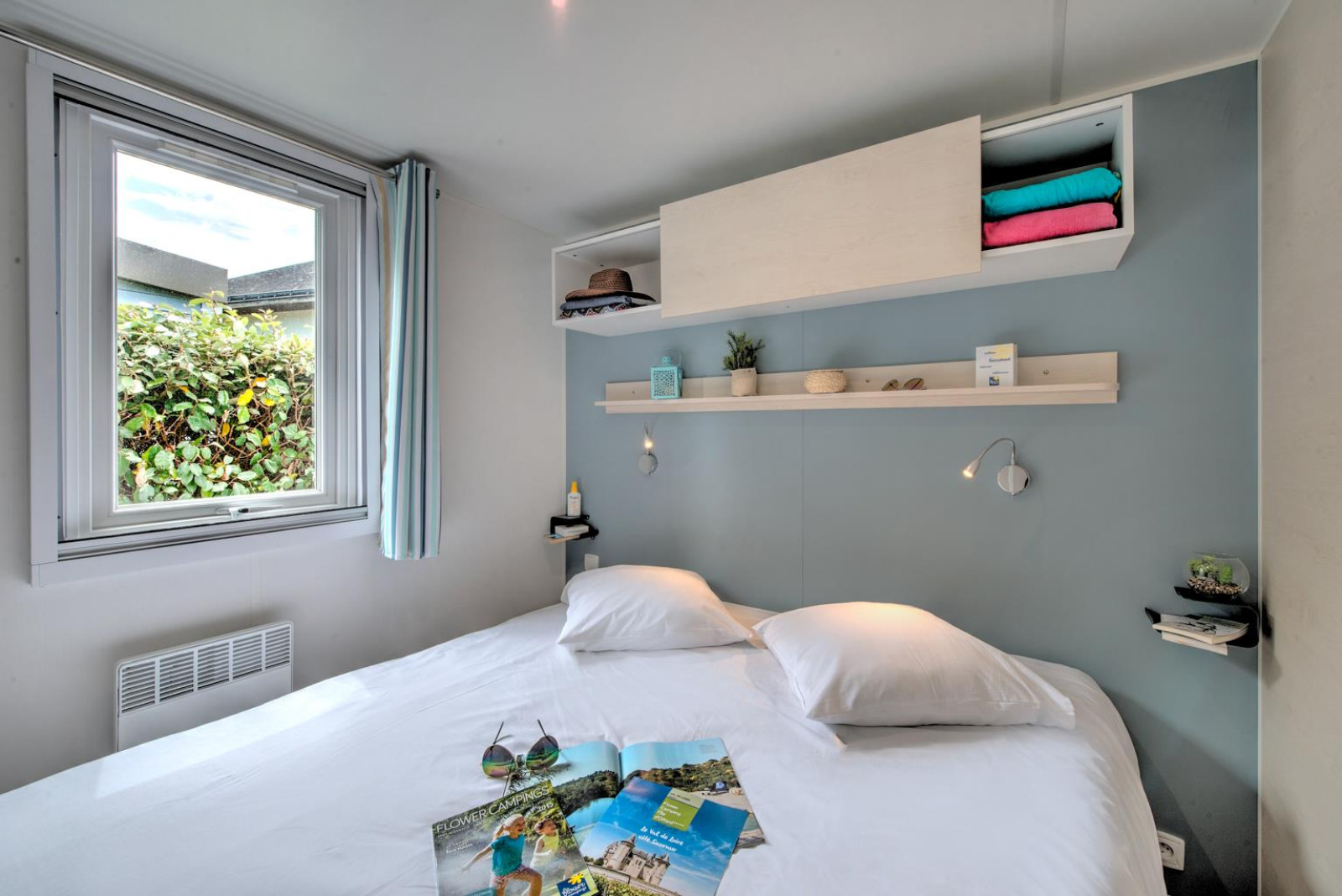 Moderne glamping-accommodatie met tweepersoonsbed, raam, open planken en gezellige, zachte inrichting.