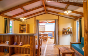 Vista interior de una acogedora cabaña glamping con muebles de madera, ventanas abiertas y terraza exterior.