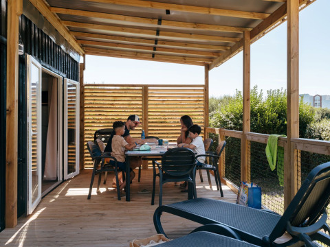 Family eating together on a covered wooden terrace at Flower Camping Le Bois d'Amour holiday park in Brittany, France.
