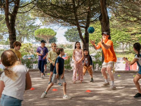 Niños juegan a la pelota con un adulto bajo los árboles en Flower Camping Le Bois d'Amour en Bretaña, Francia.